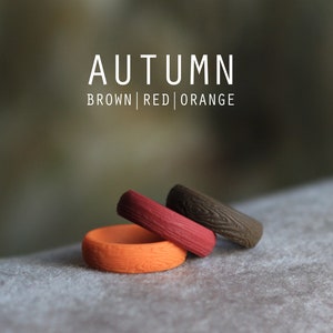 May include: Three silicone rings in brown, red, and orange with a wood grain pattern. The rings are stacked on top of each other. The text "AUTUMN BROWN | RED | ORANGE" is above the rings.