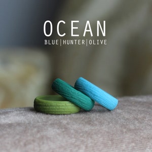 May include: Three silicone rings in shades of blue, green, and olive green. The rings have a wood grain texture. The text "OCEAN BLUE|HUNTER|OLIVE" is displayed above the rings.