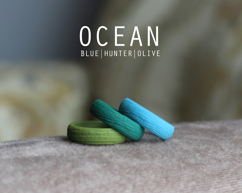 May include: Three silicone rings in shades of blue, green, and olive green. The rings have a wood grain texture. The text "OCEAN BLUE|HUNTER|OLIVE" is displayed above the rings.