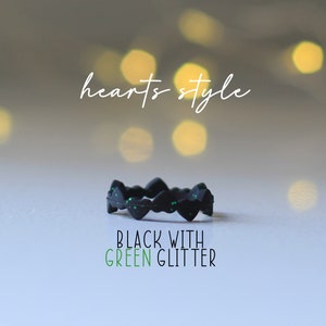 Pode incluir: Anel de silicone preto com design de cora&ccedil;&atilde;o e glitter verde. O anel est&aacute; sobre uma superf&iacute;cie branca com um fundo desfocado de luzes. O texto "hearts style" est&aacute; acima do anel e o texto "BLACK WITH GREEN GLITTER" est&aacute; abaixo do anel.