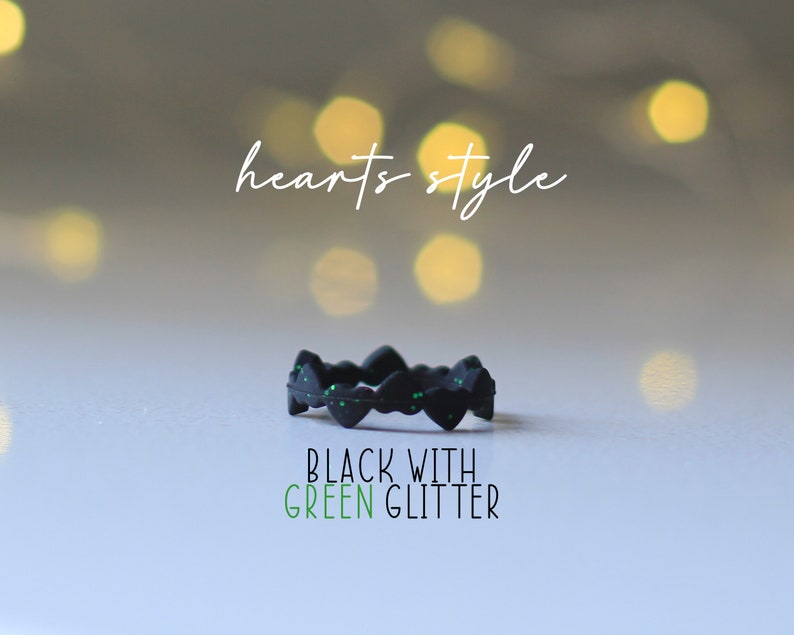 Pode incluir: Anel de silicone preto com design de cora&ccedil;&atilde;o e glitter verde. O anel est&aacute; sobre uma superf&iacute;cie branca com um fundo desfocado de luzes. O texto "hearts style" est&aacute; acima do anel e o texto "BLACK WITH GREEN GLITTER" est&aacute; abaixo do anel.
