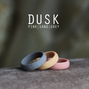 May include: Three silicone rings in pink, sand, and gray colors. The rings are stacked on top of each other. The text "DUSK PINK | SAND | GREY" is displayed above the rings.