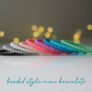 May include: A selection of beaded mini bracelets in a range of colours, such as black, white, pink, purple, and teal. The bracelets are displayed in a row, highlighting their bright colours and simple design. The text "beaded style mini bracelets" is at the bottom.