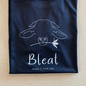 Könnte beinhalten: Marineblaues T-Shirt mit weißer Umrisszeichnung eines Schafs, das eine Blume hält. Das Wort "Bleat" ist darunter gedruckt, mit dem Satz "means I love you" darunter.