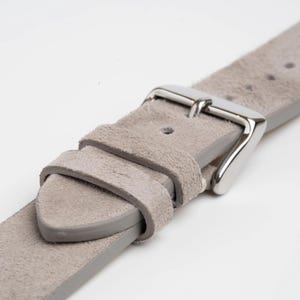 Peut inclure: Gros plan sur un bracelet de montre gris clair avec une boucle argentée. Le bracelet est fait d'un matériau semblable à du daim et présente un design classique. La boucle est brillante et contraste avec la finition mate du bracelet.