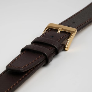 Peut inclure: Un bracelet de montre en cuir marron foncé présentant une surface texturée avec des surpiqûres marron assorties sur les bords. Le bracelet comprend une boucle en métal doré poli et deux passants en cuir coulissants.
