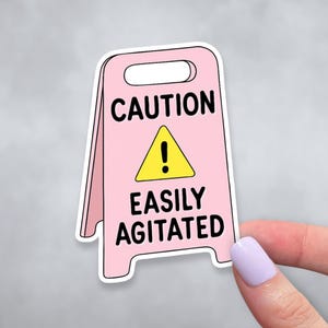 May include: A pink sticker in the shape of a caution sign with the words "CAUTION EASILY AGITATED" in black. The sign has a yellow triangle with an exclamation point. The sticker is held by a hand with a light purple nail polish.