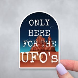 May include: A sticker with a dark blue background and white text that reads "Only Here For The UFO's". The sticker features a red rock formation against a starry night sky.