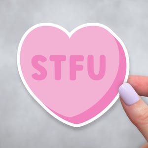 May include: A pink heart-shaped sticker with the letters "STFU" in a lighter pink font. The sticker has a white border and is held by a person with a light purple painted fingernail.
