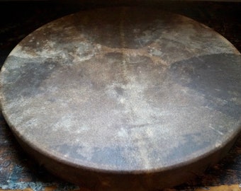 May include: A round, antique drum with a worn, textured leather surface. The drum's color is a mix of browns and grays, with darker areas suggesting age and use. The circular shape and aged appearance suggest a vintage musical instrument.