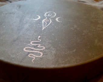 May include: A grey, round drum with a white design of a woman with a crescent moon on each side and a snake design below. The drum has a speckled surface.