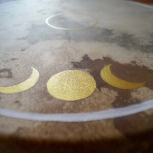 May include: A brown leather drum with a white rim and gold painted crescent moons and a full moon.