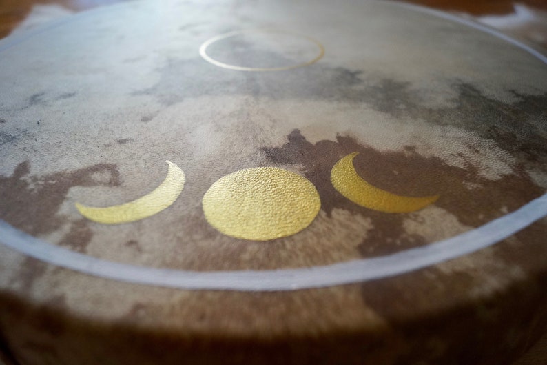 May include: A brown leather drum with a white rim and gold painted crescent moons and a full moon.
