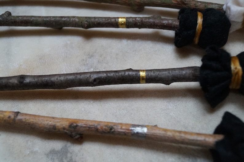 May include: Three wooden sticks with black fabric wrapped around the ends and gold tape accents. The sticks are arranged in a row on a light beige surface.