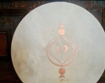 May include: A white drum with a copper-coloured design of a geometric pattern with circles and crescents. The design is centred on the drum and extends to the top and bottom.