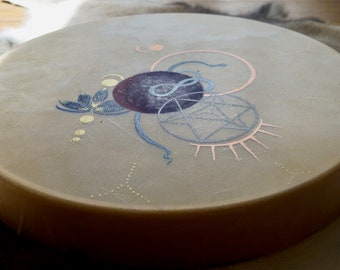 May include: A beige drum with a painted design featuring a snake, a star, a circle, and a flower. The design is in shades of blue, gold, and pink.
