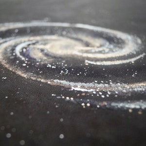 May include: A close-up of a black surface with a white and gold glitter swirl design resembling a galaxy. The design is reminiscent of a spiral galaxy with a bright center.