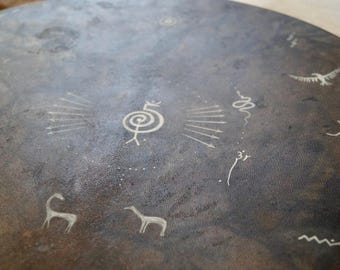 May include: A close-up of a dark gray circular surface with white markings. The markings include a spiral, arrows, a snake, a bird, and a stick figure. The surface appears to be made of stone or metal.
