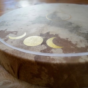 May include: A brown leather drum with a white border and gold moon phases painted on the surface.
