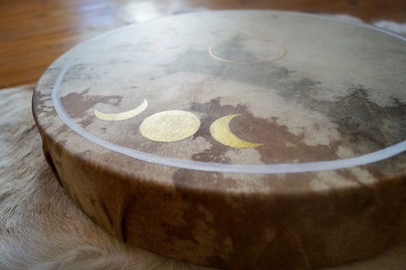May include: A brown leather drum with a white border and gold moon phases painted on the surface.