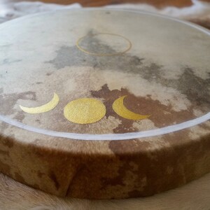 May include: A brown leather drum with a white border and gold crescent moon and sun designs.