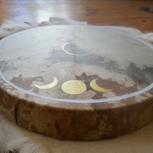 May include: A brown leather drum with a white rim and gold moon phases painted on the surface.