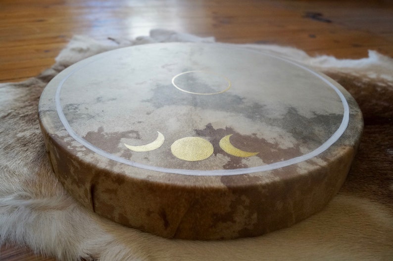 May include: A brown leather drum with a white rim and gold moon phases painted on the surface.
