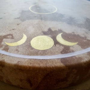 May include: A brown leather drum with a white painted circle around the edge. The center of the drum has a gold painted circle and three crescent moons.