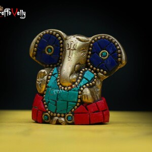 Puede incluir: Una peque&ntilde;a estatua dorada de Ganesha, el dios hind&uacute; de los comienzos, con detalles en mosaico azul y verde. La estatua est&aacute; sentada sobre una base roja y dorada.