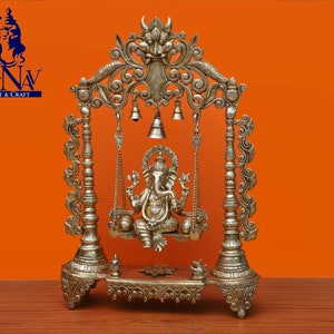 May include: A silver-coloured metal swing set with intricate carvings and bells, featuring a seated Ganesha statue. The swing set is on a wooden surface with a brown background.