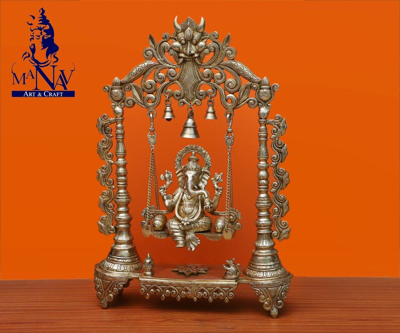 May include: A silver-coloured metal swing set with intricate carvings and bells, featuring a seated Ganesha statue. The swing set is on a wooden surface with a brown background.