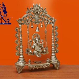 May include: Silver-toned metal statue of Ganesha, the Hindu god of wisdom and prosperity, sitting on a swing. The swing is decorated with intricate carvings and bells. The statue is on a base with a decorative design.