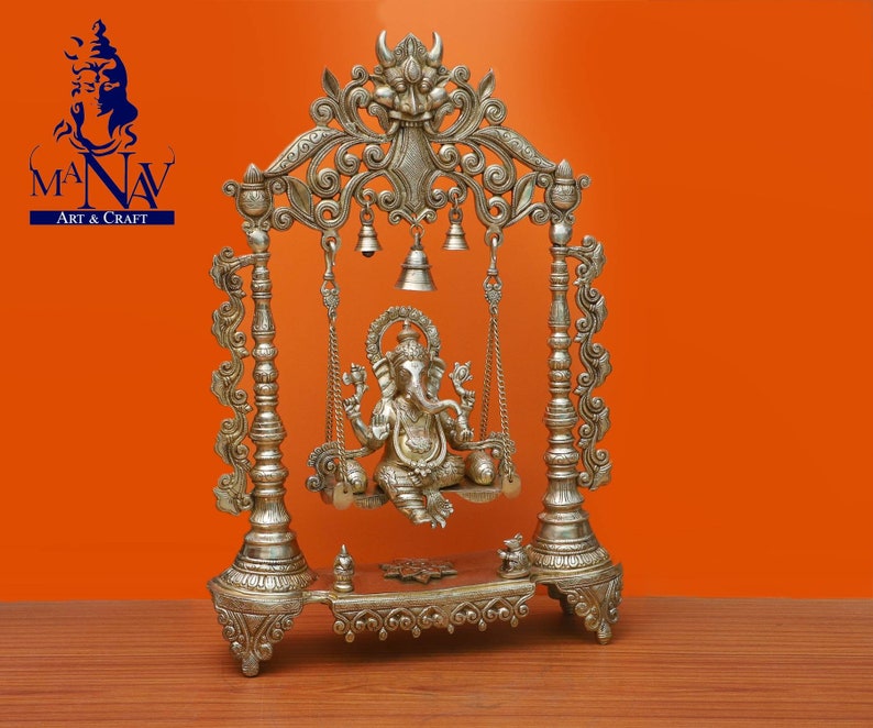 May include: Silver-toned metal statue of Ganesha, the Hindu god of wisdom and prosperity, sitting on a swing. The swing is decorated with intricate carvings and bells. The statue is on a base with a decorative design.