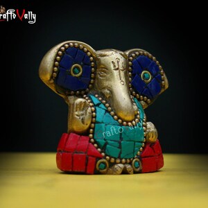 Puede incluir: Una estatua dorada de Ganesha con detalles en mosaico rojo, azul y verde. La estatua est&aacute; sentada sobre una superficie amarilla.