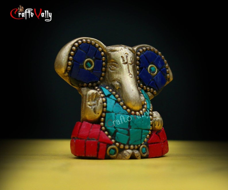 Puede incluir: Una estatua dorada de Ganesha con detalles en mosaico rojo, azul y verde. La estatua est&aacute; sentada sobre una superficie amarilla.
