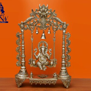 May include: A silver-coloured metal sculpture of the Hindu deity Ganesha, sitting on a swing. The sculpture is ornate and detailed, with intricate carvings and embellishments. The swing is suspended from a decorative archway, which is also adorned with bells.