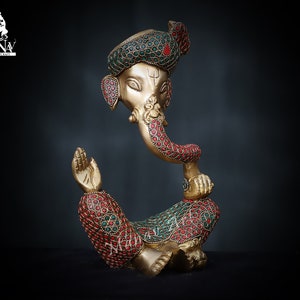 May include: A gold-colored statue of Ganesha, the Hindu god of wisdom and prosperity, with red and green gemstone accents. The statue is in a seated position with the god's trunk in his mouth.