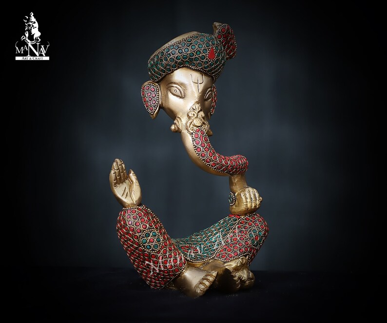 May include: A gold-colored statue of Ganesha, the Hindu god of wisdom and prosperity, with red and green gemstone accents. The statue is in a seated position with the god's trunk in his mouth.