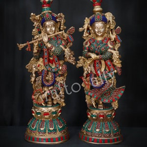 May include: A pair of gold-toned statues of Hindu deities, Radha and Krishna, with intricate details and colourful gemstone accents. The statues are standing on ornate bases.