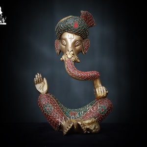 May include: A gold-colored statue of Ganesha, the Hindu god of beginnings, prosperity, and good fortune. The statue is decorated with red and green gemstones and is sitting in a meditative pose.