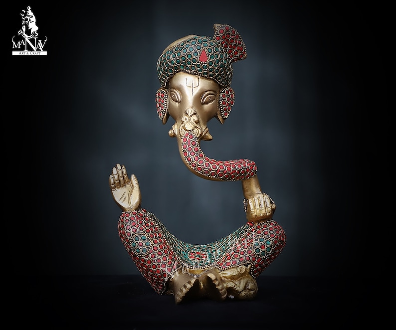 May include: A gold-colored statue of Ganesha, the Hindu god of beginnings, prosperity, and good fortune. The statue is decorated with red and green gemstones and is sitting in a meditative pose.