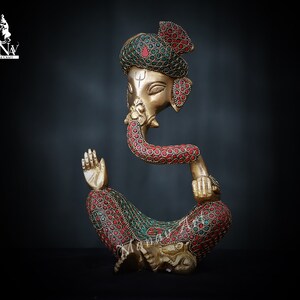 May include: A gold-colored metal statue of Ganesha, the Hindu god of wisdom and prosperity. The statue is decorated with red and green gemstones. Ganesha is depicted sitting with his trunk in his mouth and his right hand raised in a gesture of blessing.