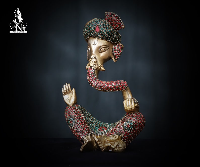 May include: A gold-colored metal statue of Ganesha, the Hindu god of wisdom and prosperity. The statue is decorated with red and green gemstones. Ganesha is depicted sitting with his trunk in his mouth and his right hand raised in a gesture of blessing.