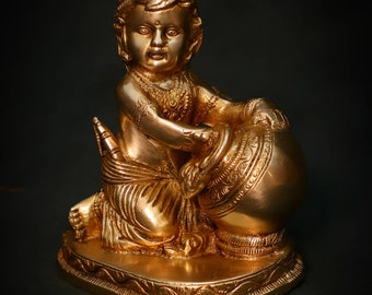 May include: A gold-coloured metal statue of a young boy holding a pot. The boy is wearing a crown and a decorative garment. The statue is sitting on a heart-shaped base.