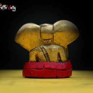 Puede incluir: Una peque&ntilde;a figura de elefante dorada con base de mosaico rojo. El elefante est&aacute; sentado con la trompa levantada y las orejas extendidas. La figura es un dise&ntilde;o tradicional indio.