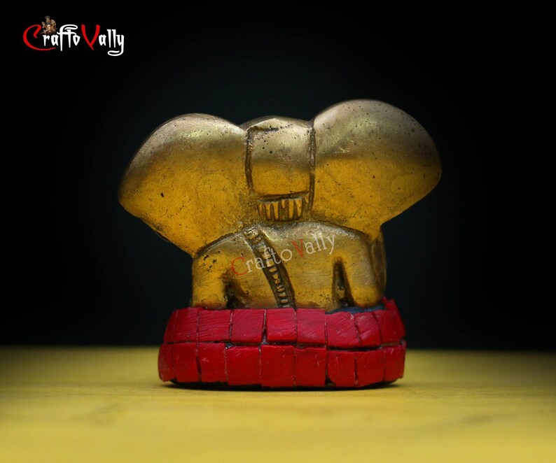 Puede incluir: Una peque&ntilde;a figura de elefante dorada con base de mosaico rojo. El elefante est&aacute; sentado con la trompa levantada y las orejas extendidas. La figura es un dise&ntilde;o tradicional indio.