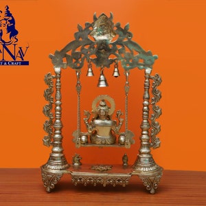 May include: A gold-coloured metal swing set with a deity figure sitting on the swing. The swing set has intricate details and is decorated with bells. The swing set is on a wooden surface.