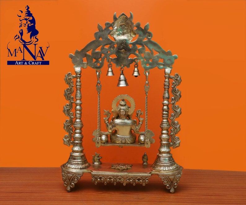 May include: A gold-coloured metal swing set with a deity figure sitting on the swing. The swing set has intricate details and is decorated with bells. The swing set is on a wooden surface.