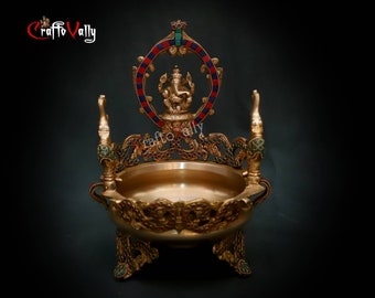 Puede incluir: Un soporte decorativo de metal dorado con una parte superior redonda y tres patas. El soporte está decorado con diseños intrincados y piedras preciosas de colores. Una pequeña estatua de la deidad hindú Ganesha se sienta en la parte superior del soporte.