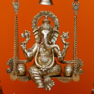 May include: A gold-coloured metal statue of Ganesha, the Hindu god of wisdom and prosperity, sitting on a swing. The statue is intricately detailed and has a traditional design.
